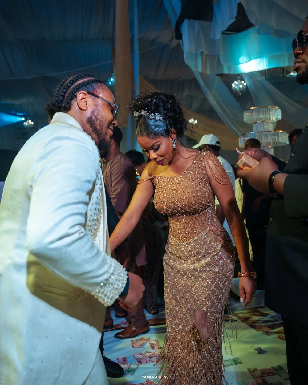 Two people dance close together at a celebration, wearing formal attire under blue lighting and draped decor.