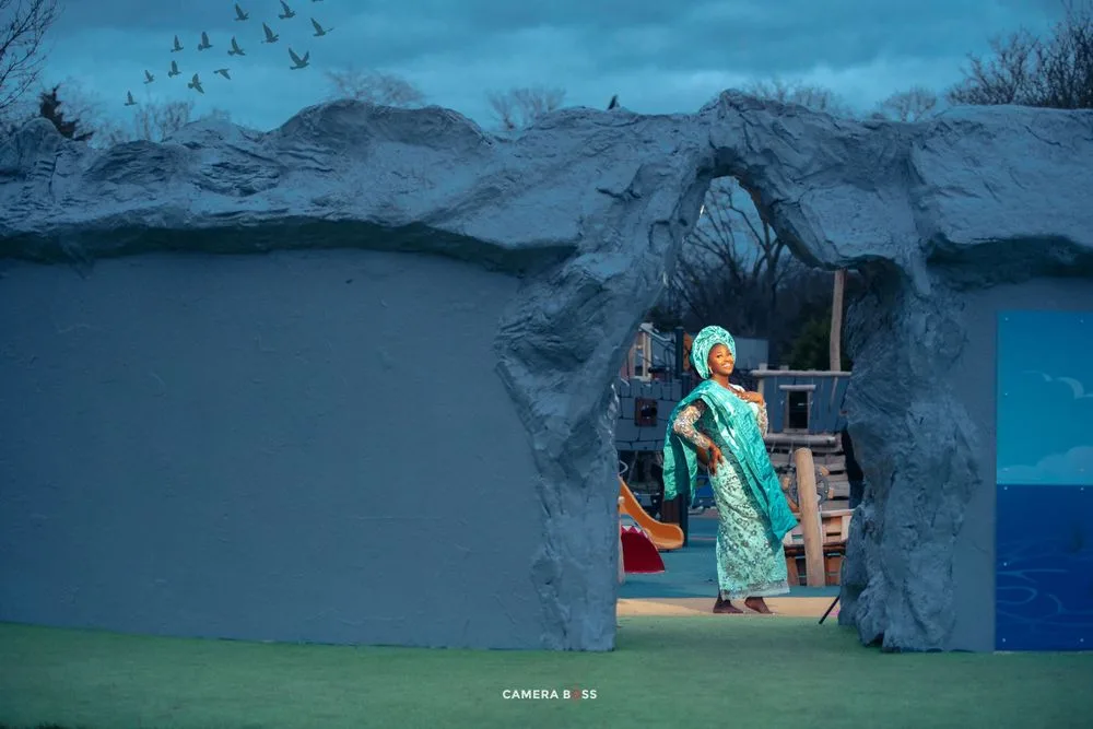 A person in a turquoise dress stands in a cave-like setting with dramatic blue lighting and mountains in the background.