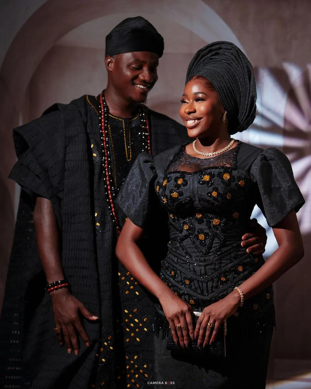 Nigerian couple in matching black traditional aso-oke outfits with gele and cap smiling together.
