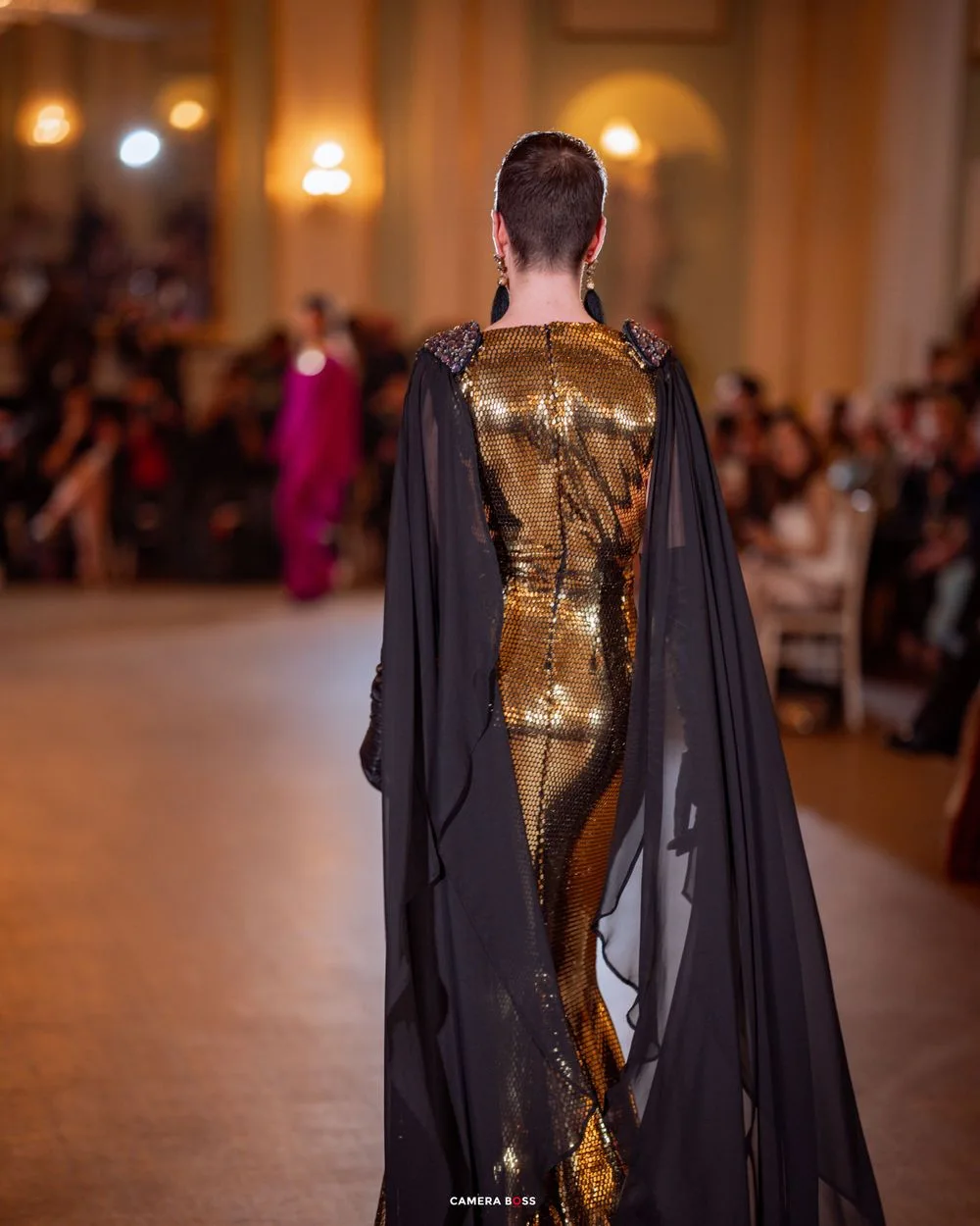 Model wearing a dramatic gold metallic gown with flowing black cape on a fashion runway with audience in background.