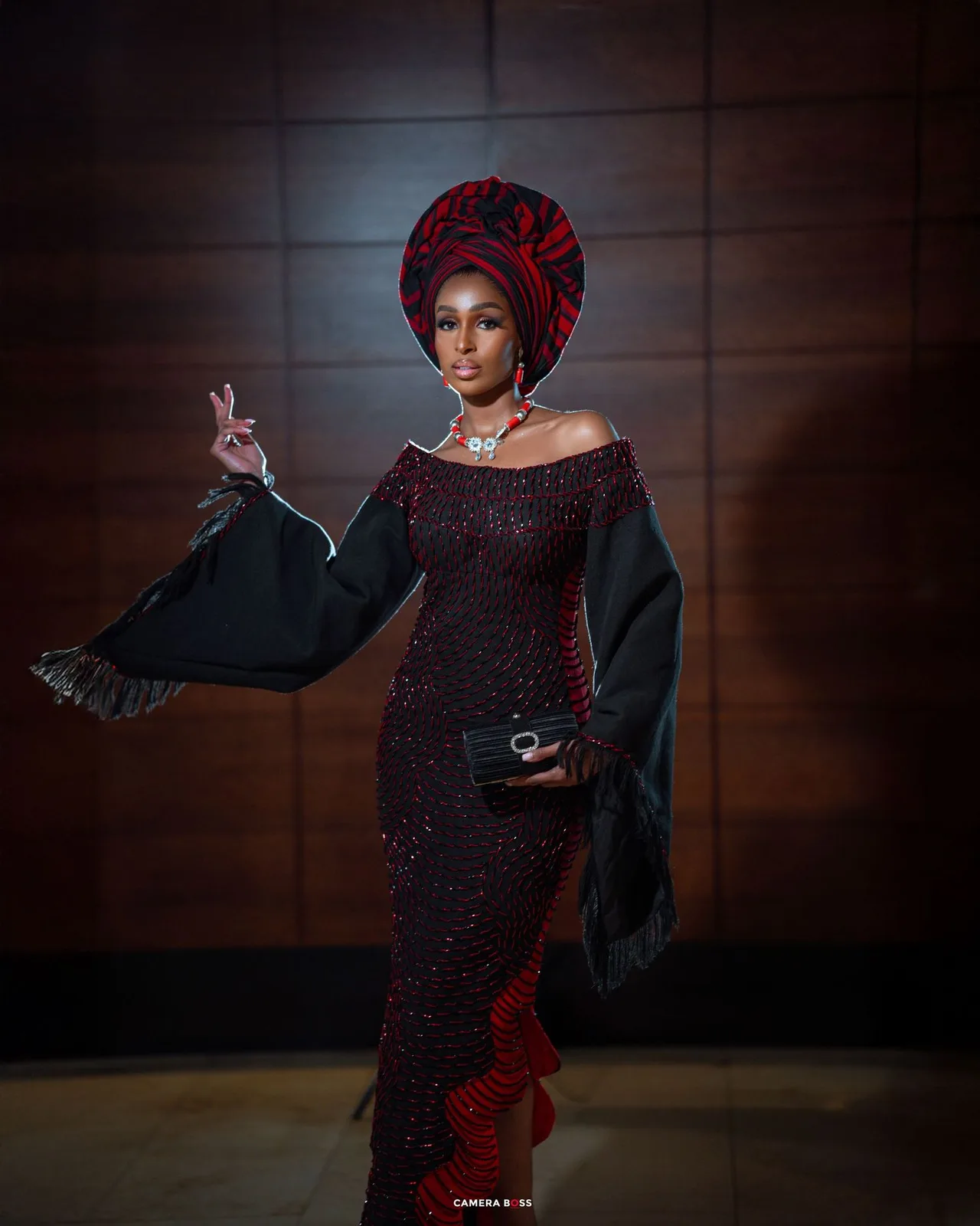 Elegant figure in a sparkly dark dress with dramatic bell sleeves and red headwrap poses dramatically against wooden paneling.