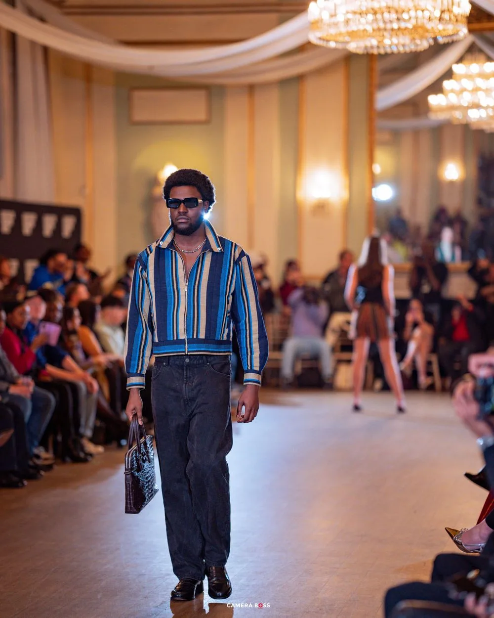 Model walks runway in striped blue shirt and dark pants during fashion show in elegant ballroom setting.