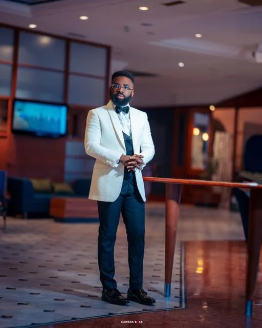 Stylishly dressed individual in white blazer and dark pants stands confidently in an elegant indoor venue with ambient lighting.