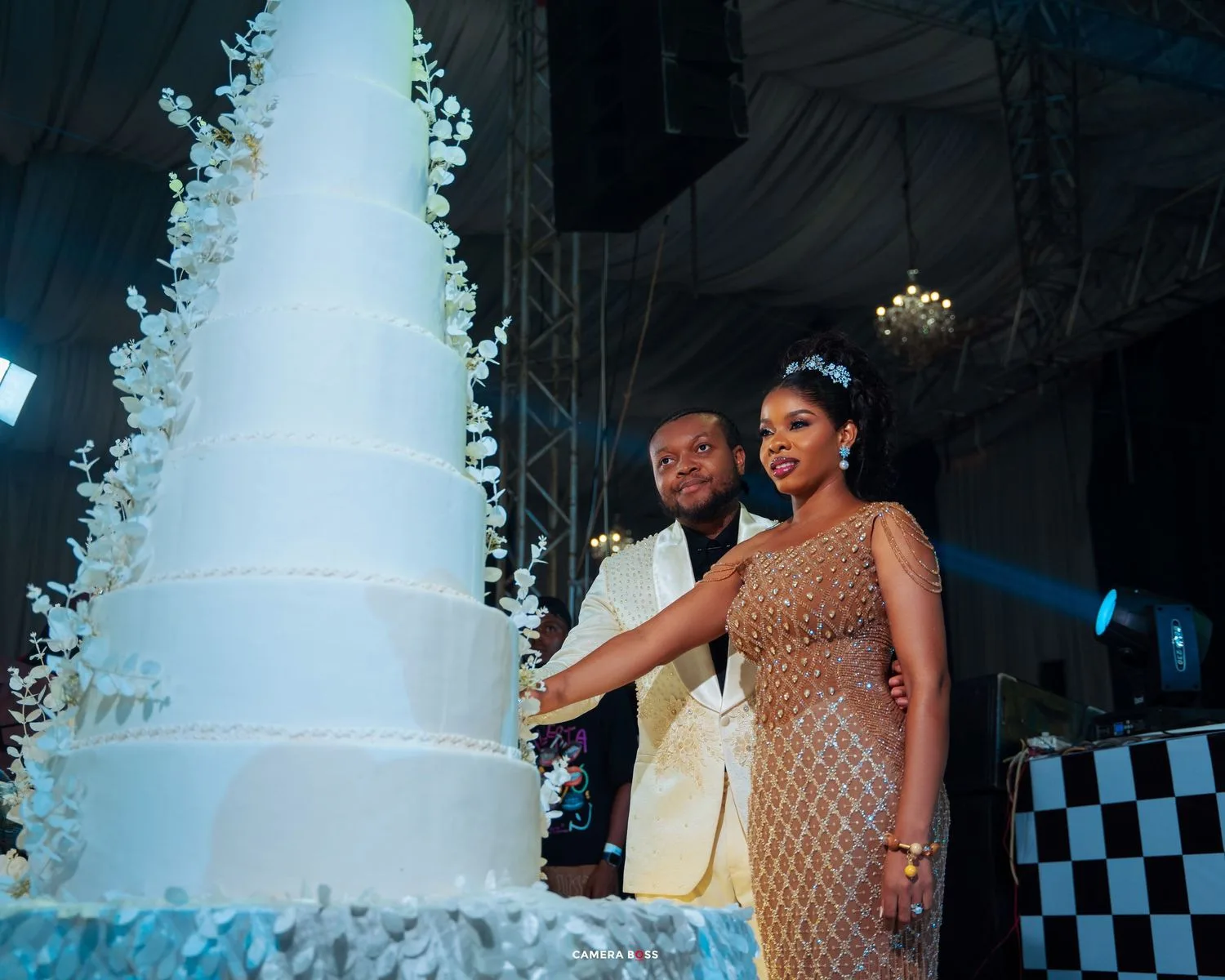 A couple cuts into a tall white wedding cake adorned with cascading greenery under dramatic blue lighting.