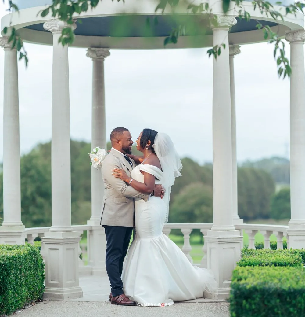 Newlywed couple embraces under an elegant white gazebo surrounded by manicured gardens and greenery.
