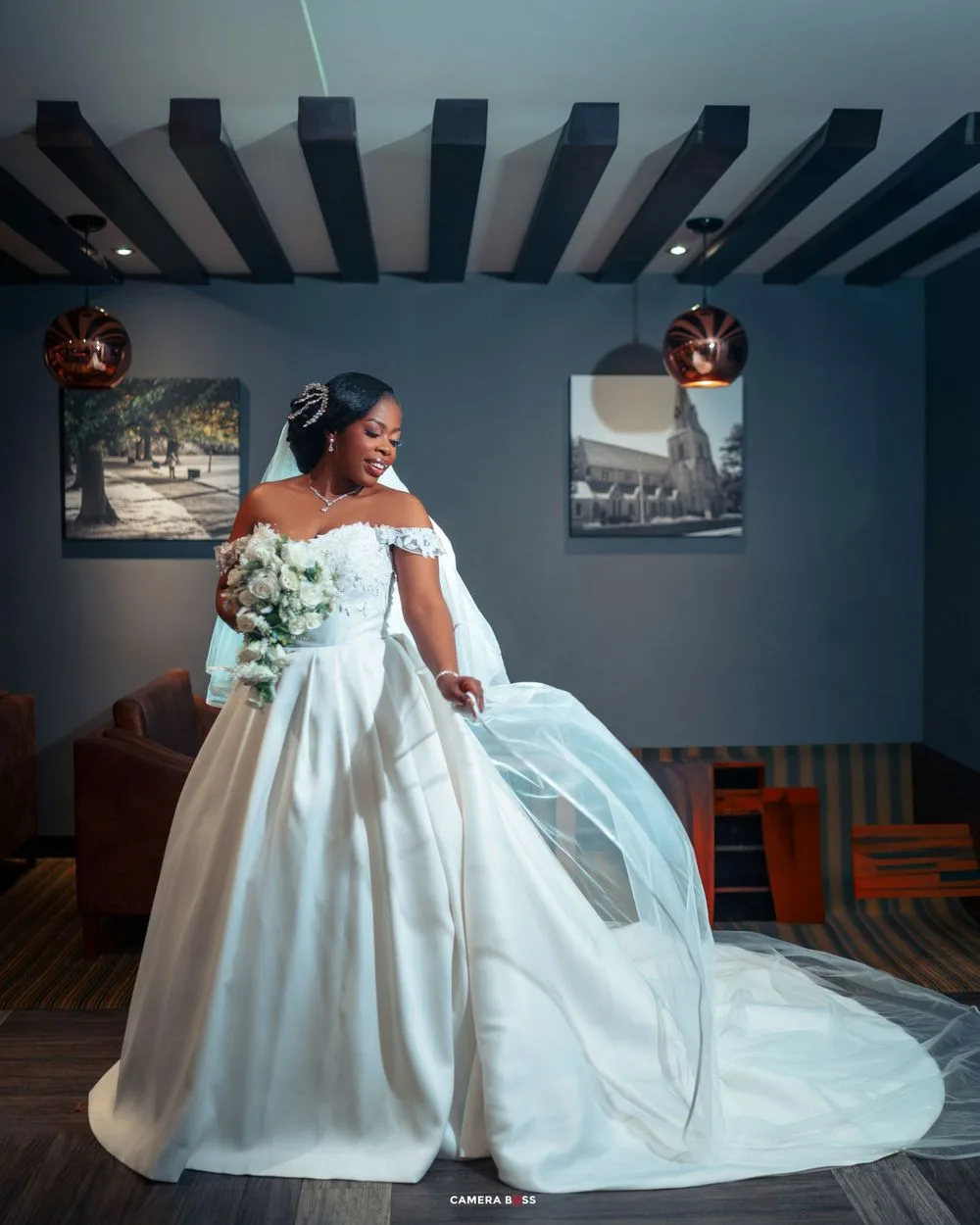 Beautiful bride in flowing off-shoulder white ball gown twirls elegantly in a room with beamed ceiling and wall art.