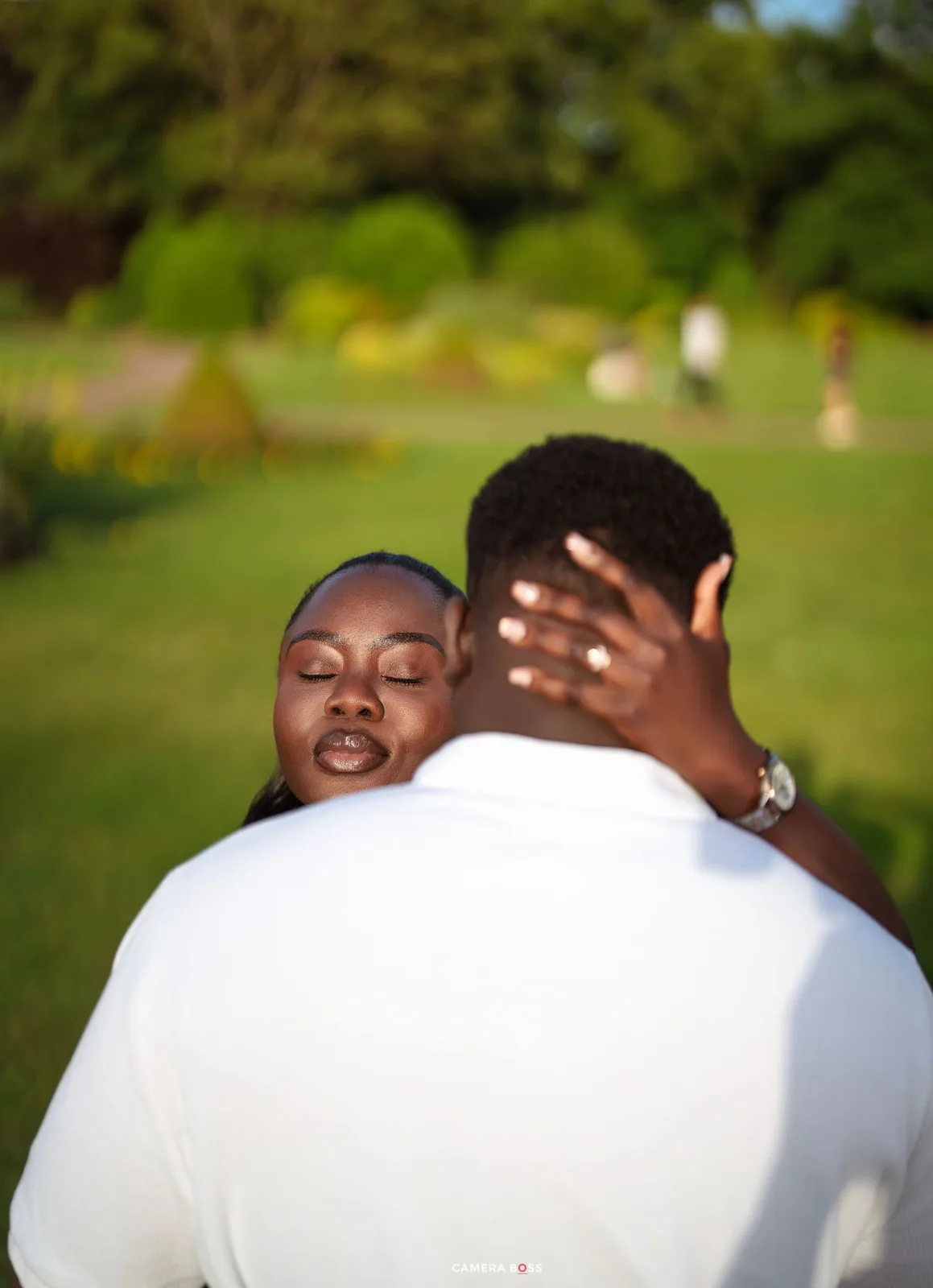 A couple embracing in a lush green park, woman resting her head on man's shoulder with eyes closed.