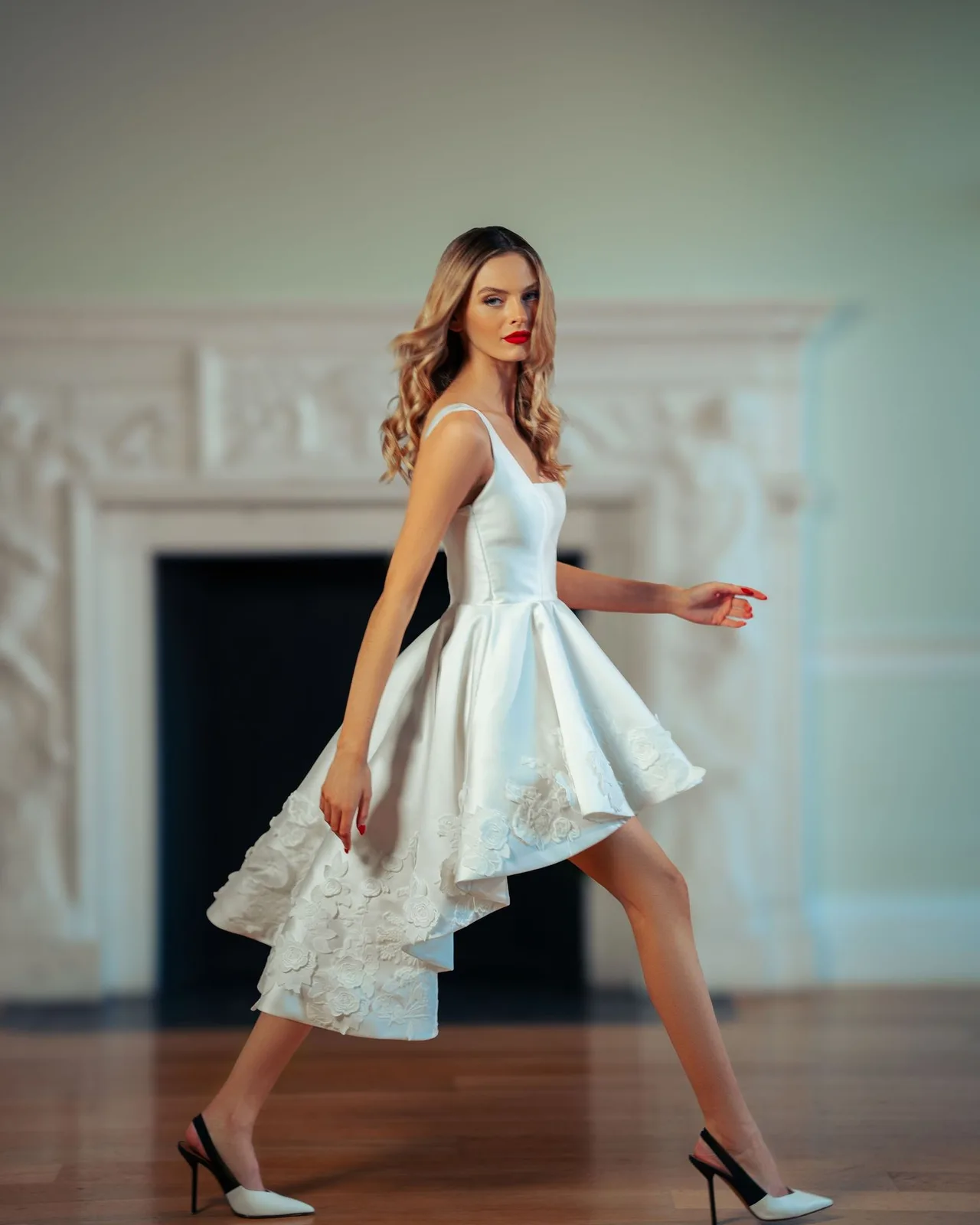Elegant white high-low dress with lace details and flowing skirt showcased in a room with ornate fireplace.