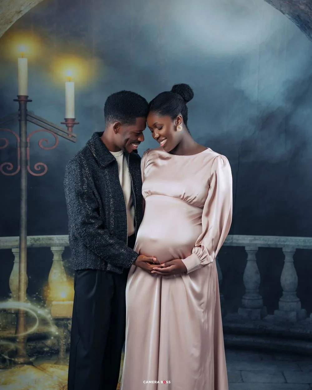 Atmospheric maternity photo with candlelit balcony, featuring couple in pink gown and dark sweater against moody backdrop.