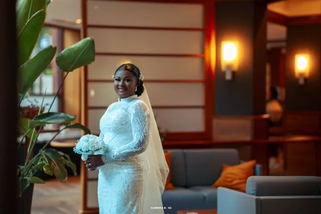 Beautiful bride in white lace gown with long sleeves and veil standing in elegant hotel lobby with warm lighting.
