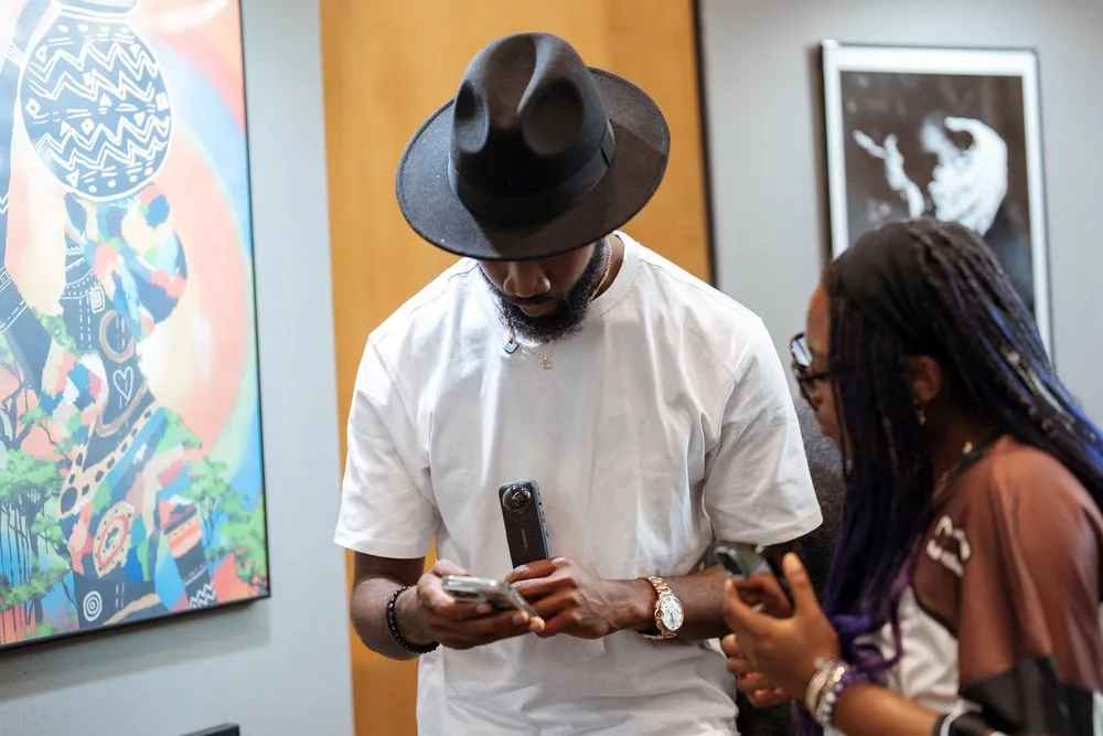 Two people in an art gallery look at a phone, one wearing a black wide-brim hat and white shirt.