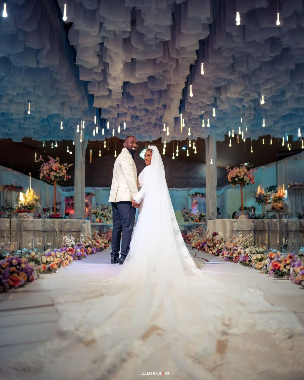 Romantic wedding scene with cascading paper cloud ceiling and hanging lights above a floral-lined venue.