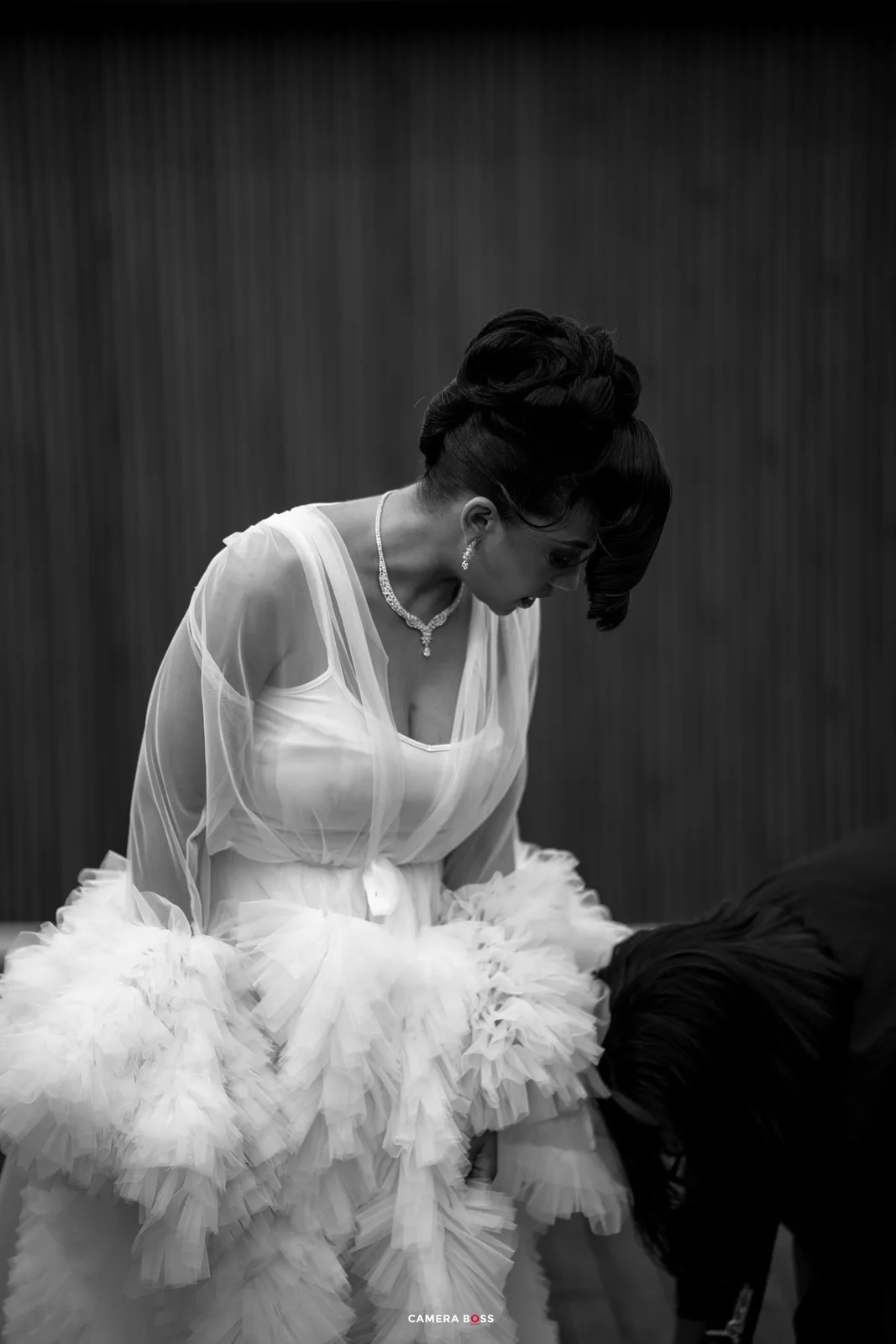 Elegant figure in white gown with sheer overlay and tulle skirt gazes down in dramatic black and white portrait.