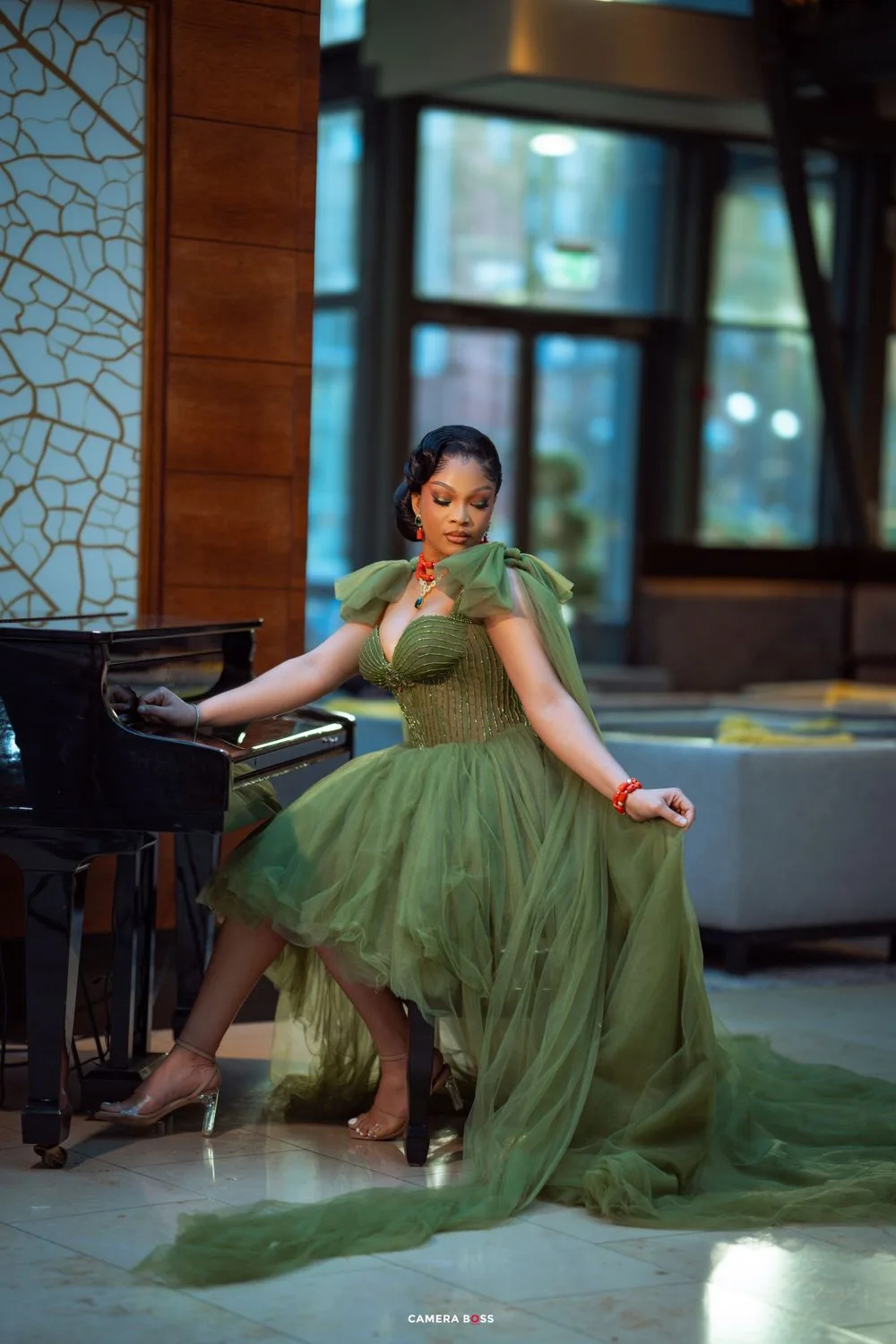 Elegant green tulle gown with train flows around model posing gracefully by grand piano in luxurious interior setting.
