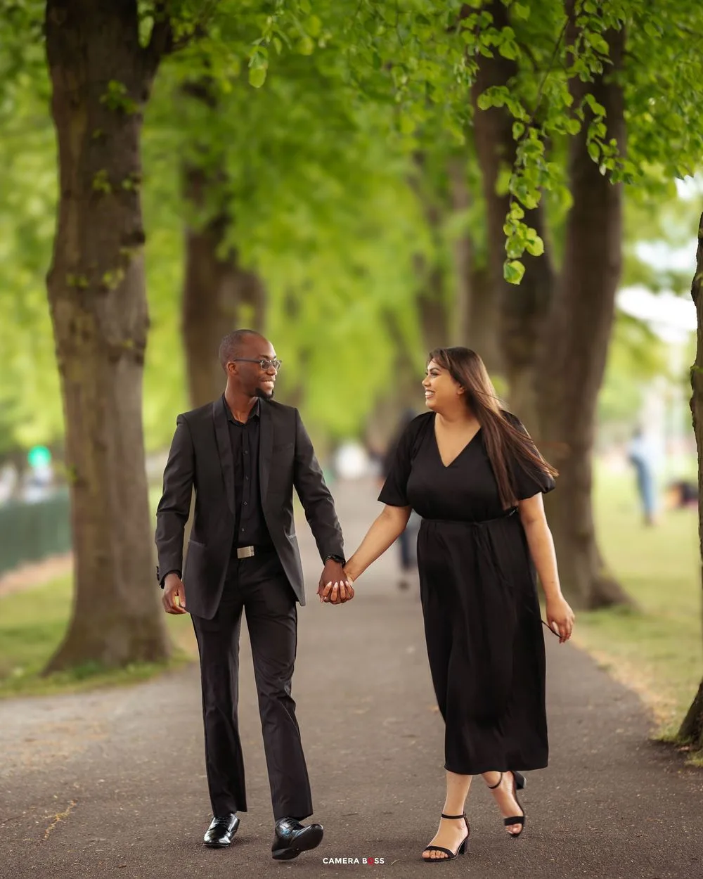 A happy couple walks hand in hand down a picturesque tree-lined pathway while wearing matching black formal attire.