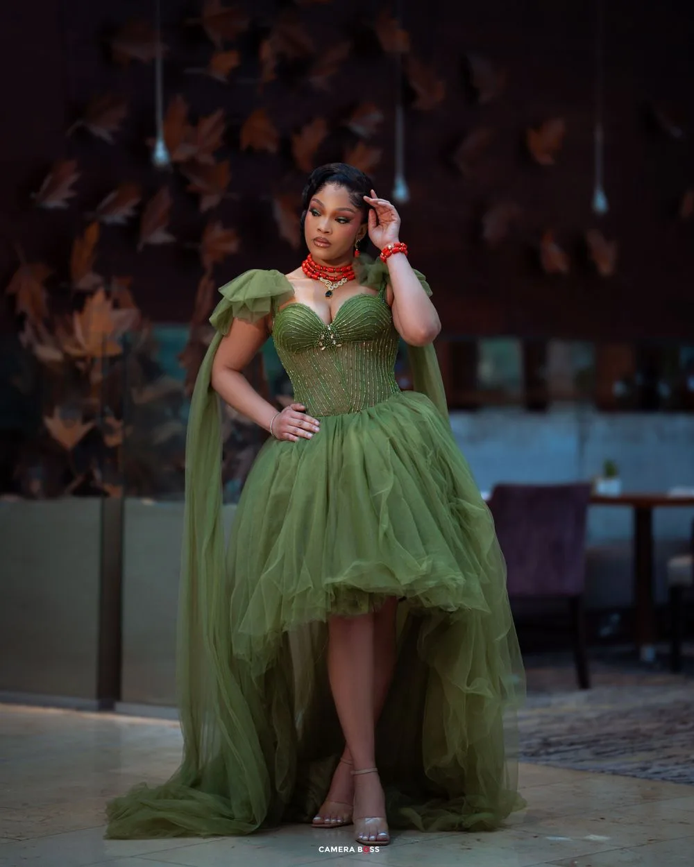 Evening gown featuring an olive green high-low tulle skirt and bodice with off-shoulder sleeves and a red necklace detail.