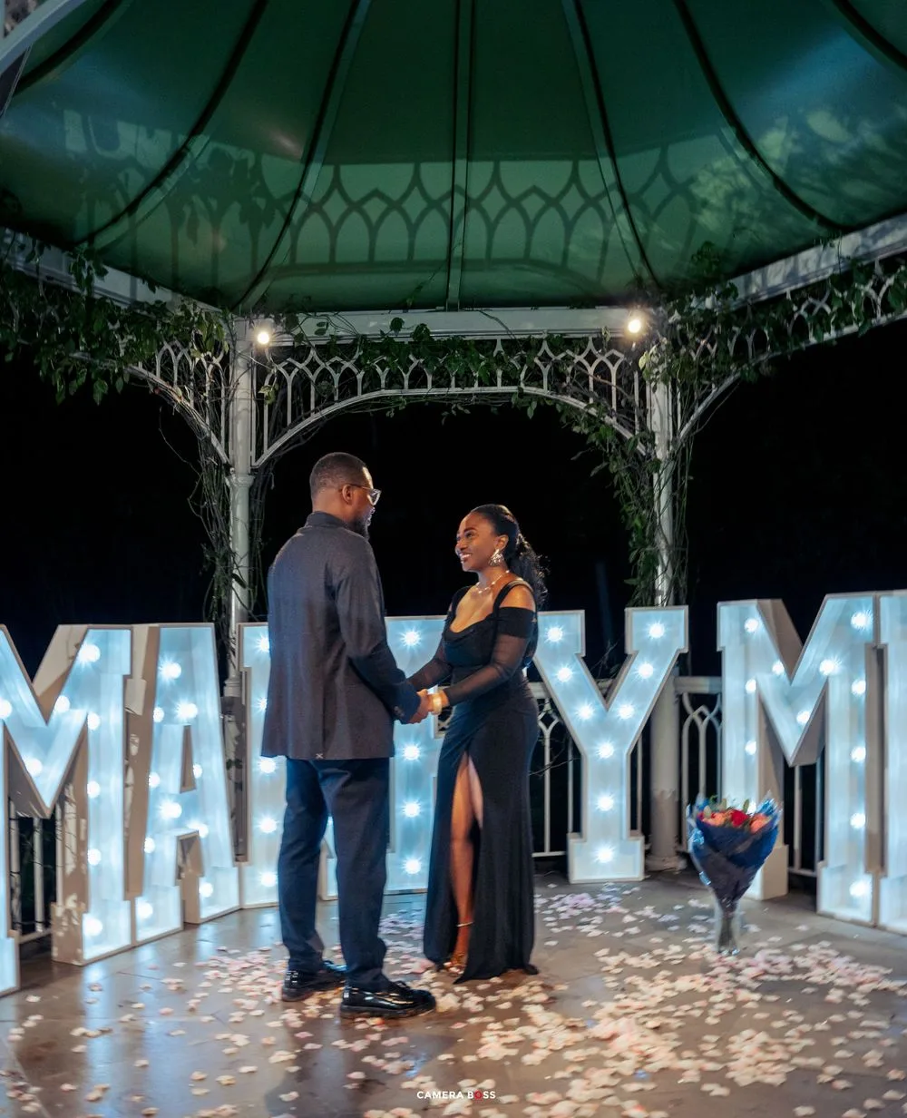 Romantic couple standing in an illuminated gazebo at night with lit 'MARRY ME' letters glowing in the background.