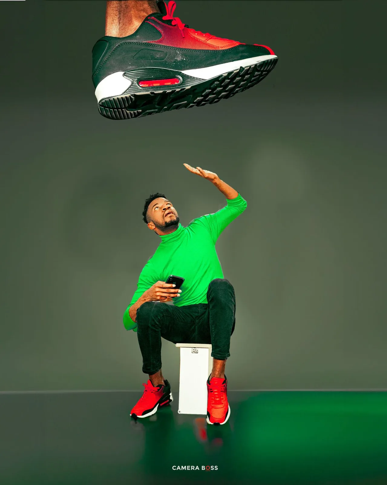 A person in green attire sits on a white box as a red sneaker floats above in a stylized studio photo.