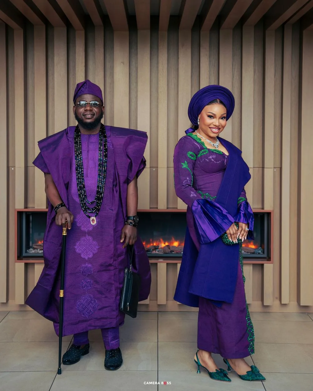 Couple wearing coordinated traditional Nigerian outfits in rich purple and blue tones standing by a fireplace.