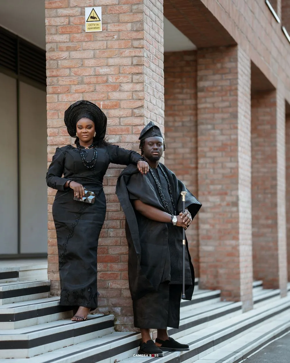 Two people in black formal attire stand elegantly on concrete steps against a brick wall backdrop.