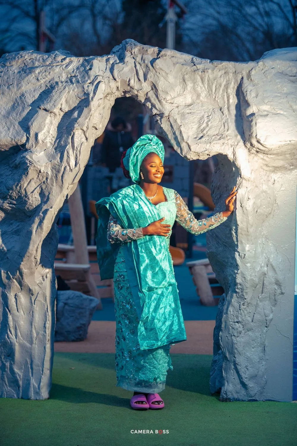 Woman in vibrant turquoise traditional African dress and headwrap poses elegantly between white stone archway at dusk.