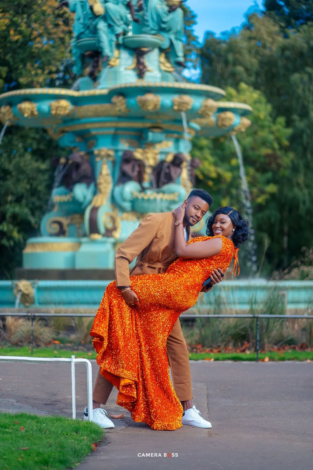 Romantic couple poses playfully by an ornate turquoise fountain, with person in orange dress and partner in tan outfit.