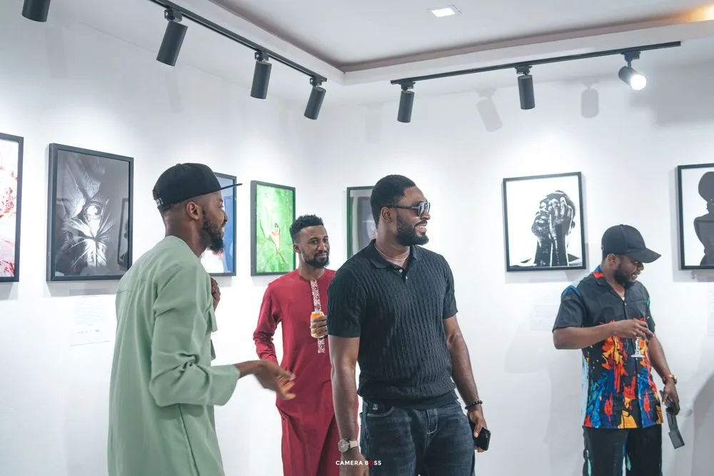 People viewing artwork displayed in a white-walled gallery with track lighting and framed photographs on the walls.