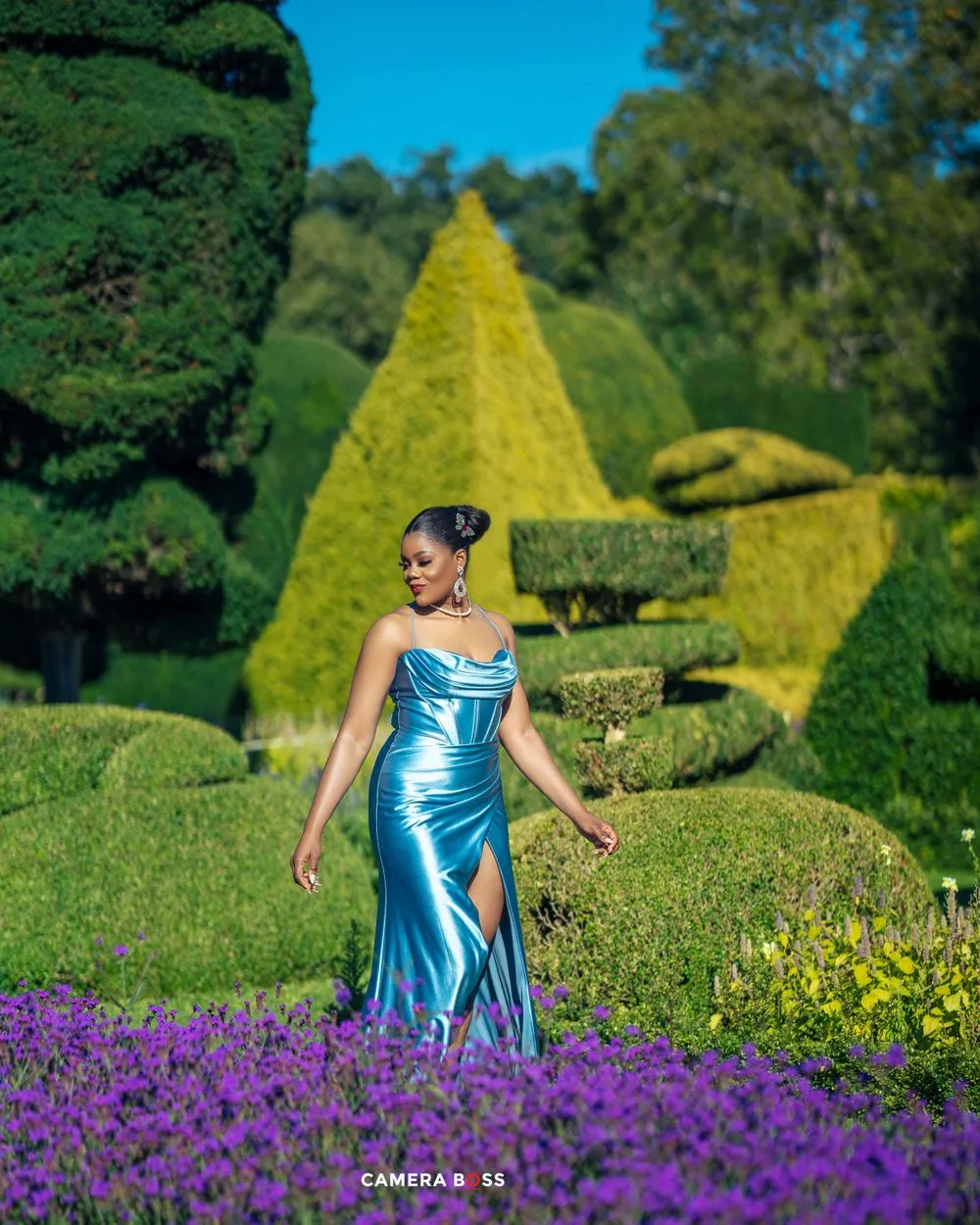 Shimmering metallic blue gown stands out against vibrant purple flowers and sculptured topiary garden on a sunny day.