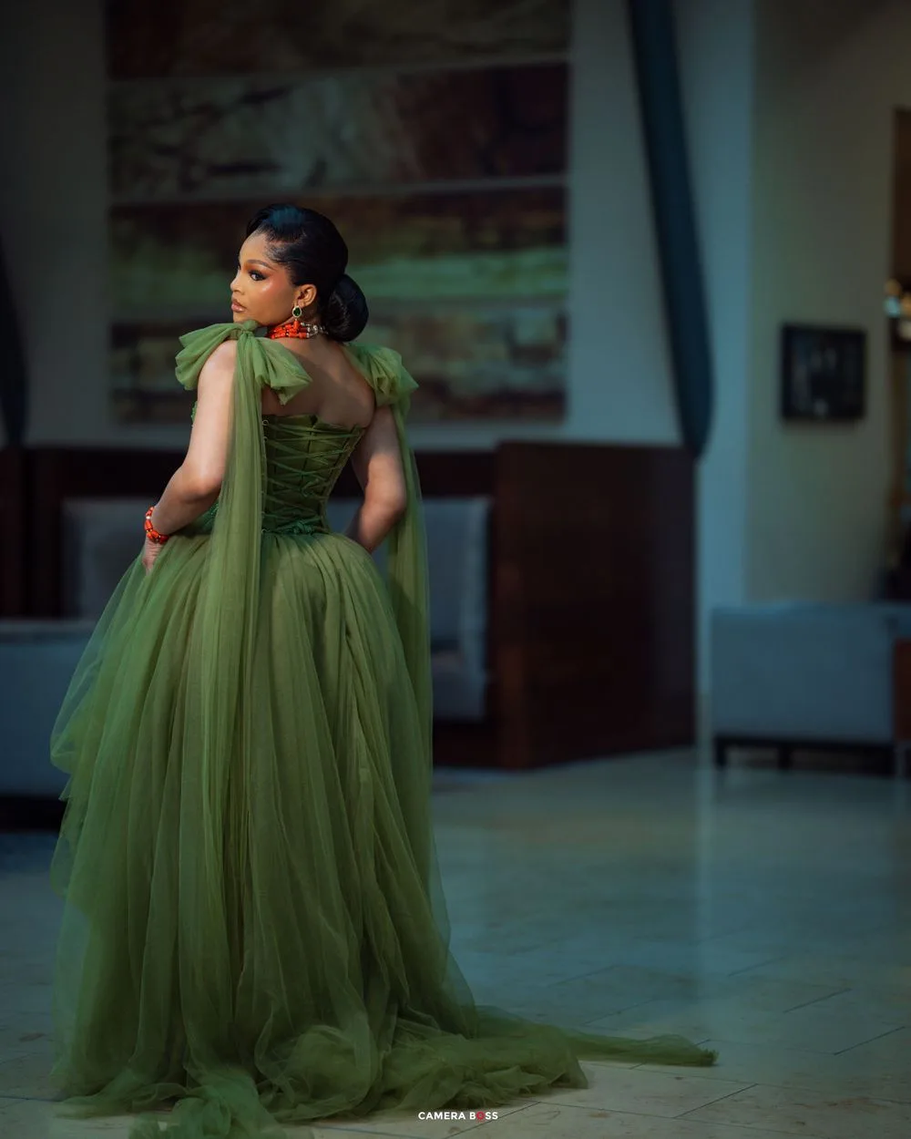 Back view of an olive green ballgown with dramatic tulle skirt and flowing cape details in dim lighting.