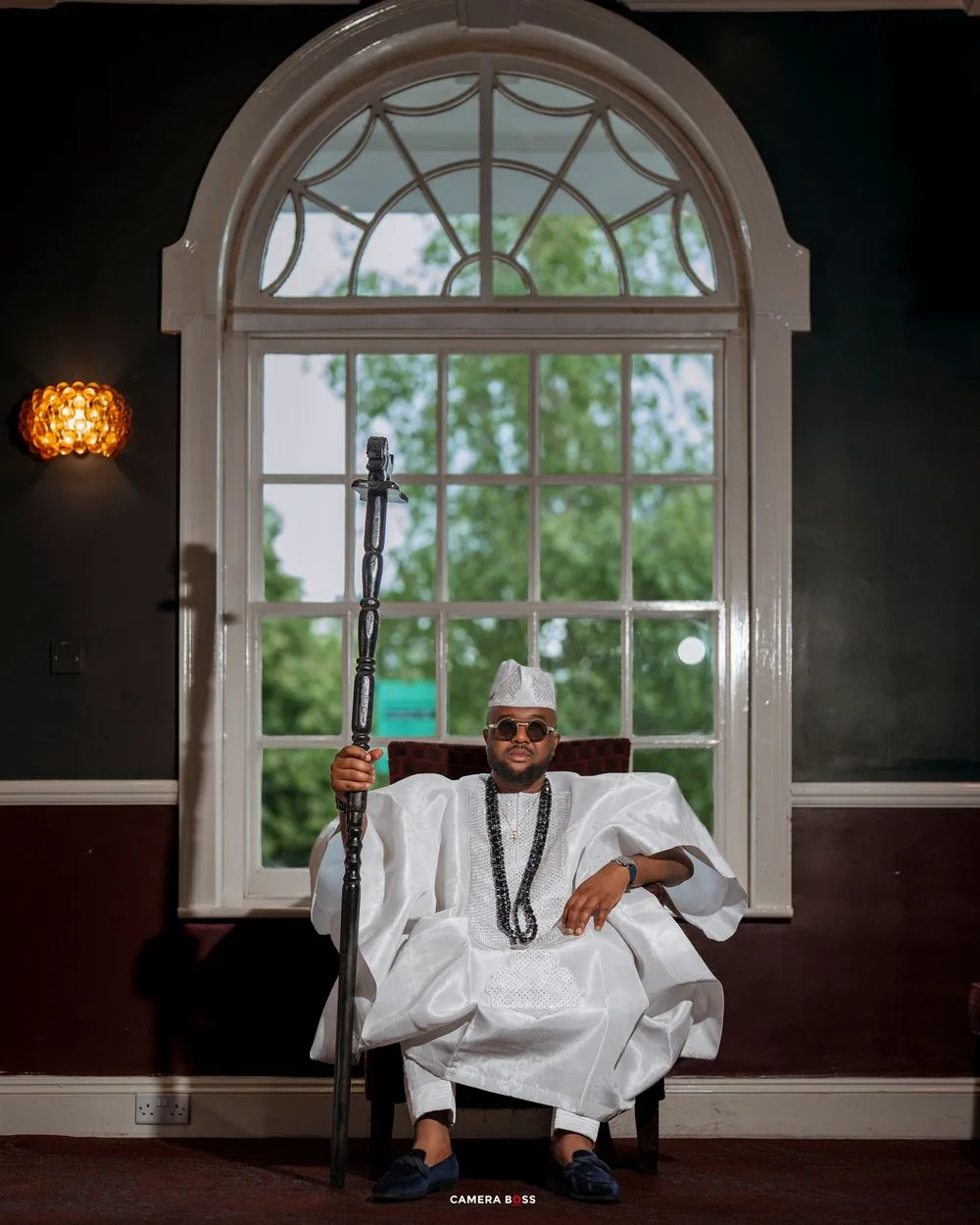 Man in white traditional Nigerian agbada attire holding a staff, seated before an arched window.