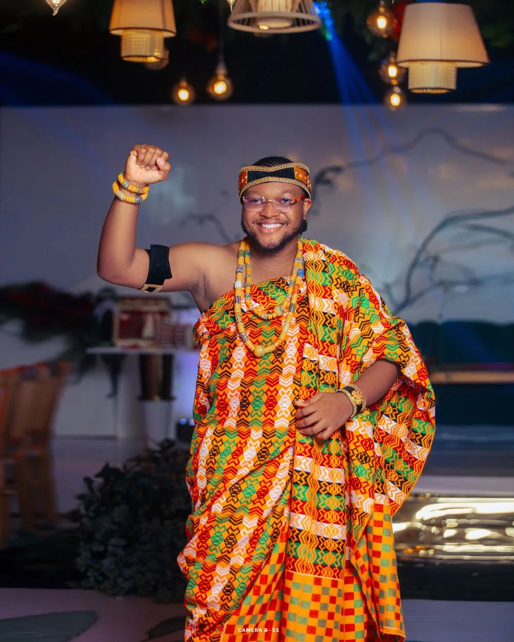 Traditional African attire in orange and green kente patterns worn during festive celebration under ambient lighting.