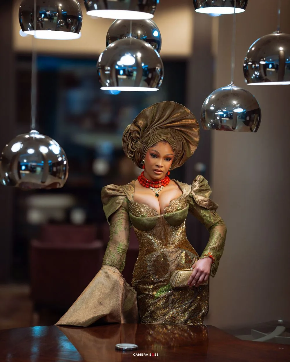 Chrome pendant lamps illuminate an elegant figure in a metallic green gown with dramatic headpiece at a dark restaurant.