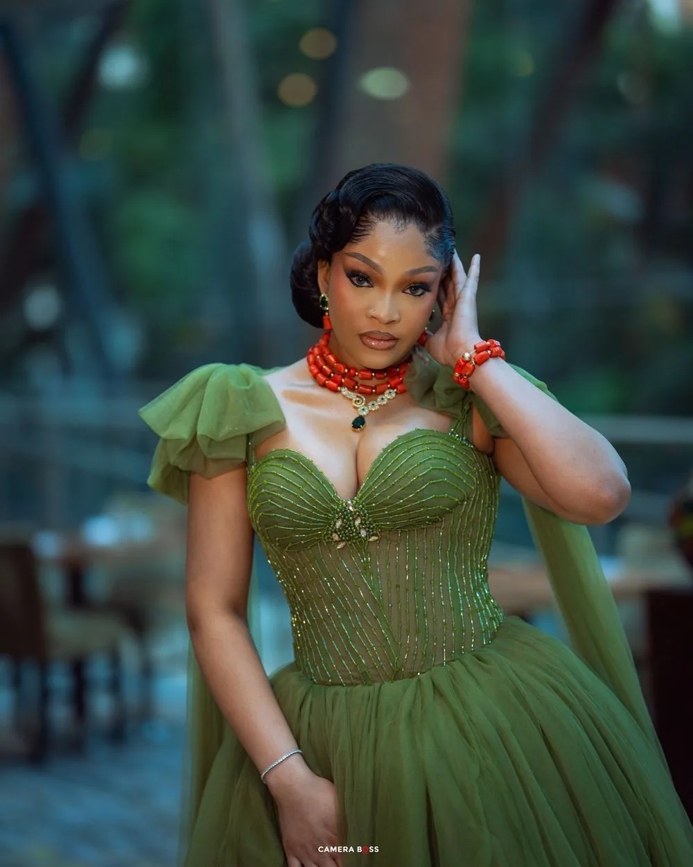 A woman in an olive green princess-style dress with ruched sleeves and embellished bodice poses against a blurred background.