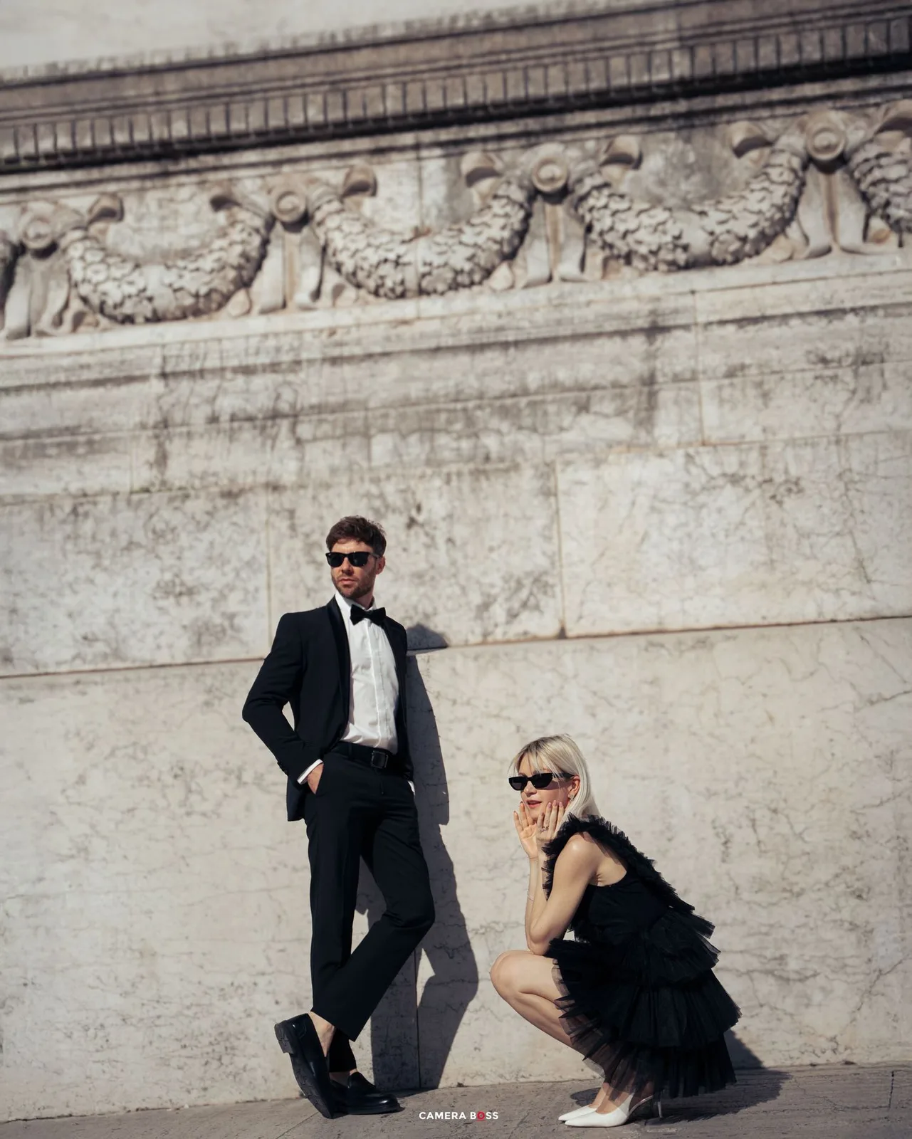 Two people in elegant black outfits pose stylishly against an ornate stone wall with classical architectural details.